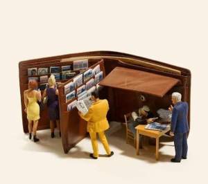 Geldbeutel ist aufgeklappt, in den Karten-Fächern stecken Miniatur-Zeitungen, die Miniaturfiguren wie an einem Zeitschriftenkiosk ansehen. 