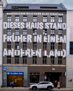 Hausfassade (vermutlich aus Ostberlin) mit Schriftzug „Dieses Haus stand früher in einem anderen Land“. Darüber: „Menschlicher Wille kann alles versetzen“.